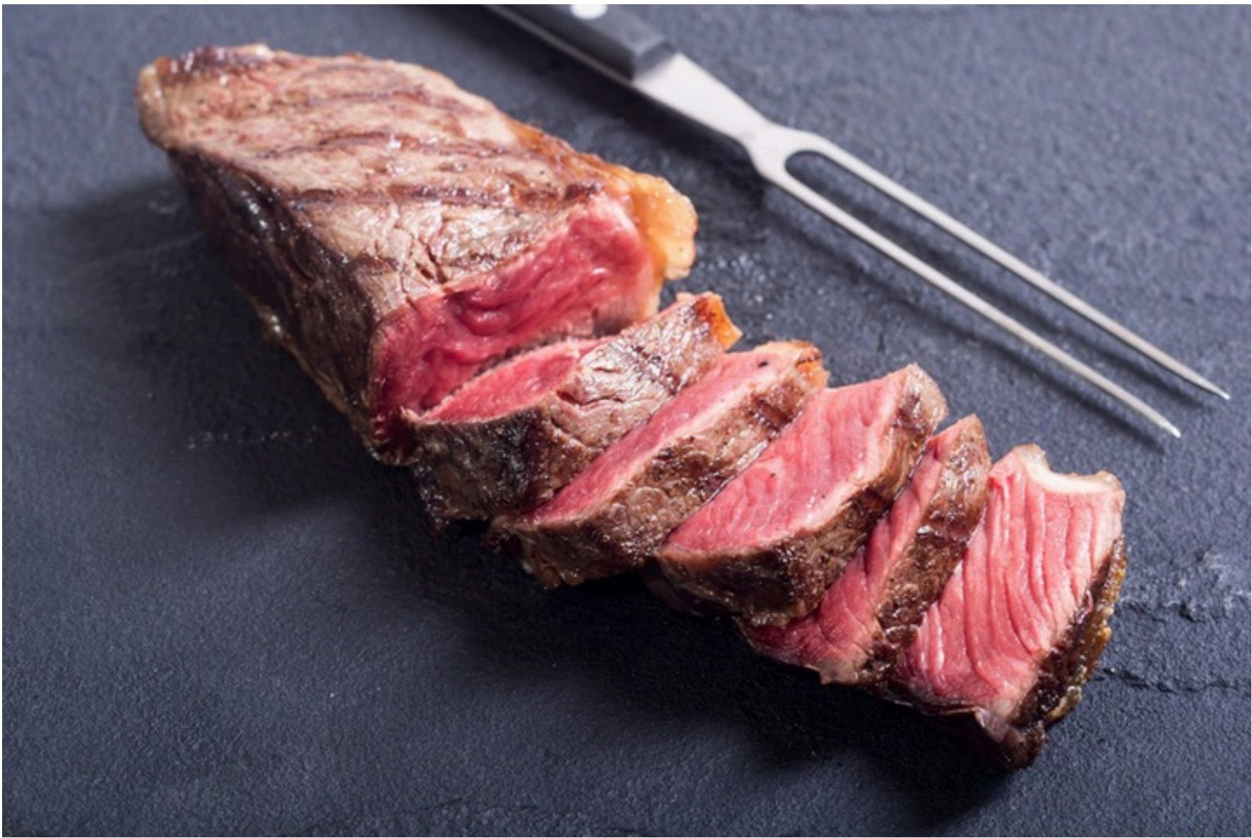 Sliced cooked steak on a dark surface with a carving fork